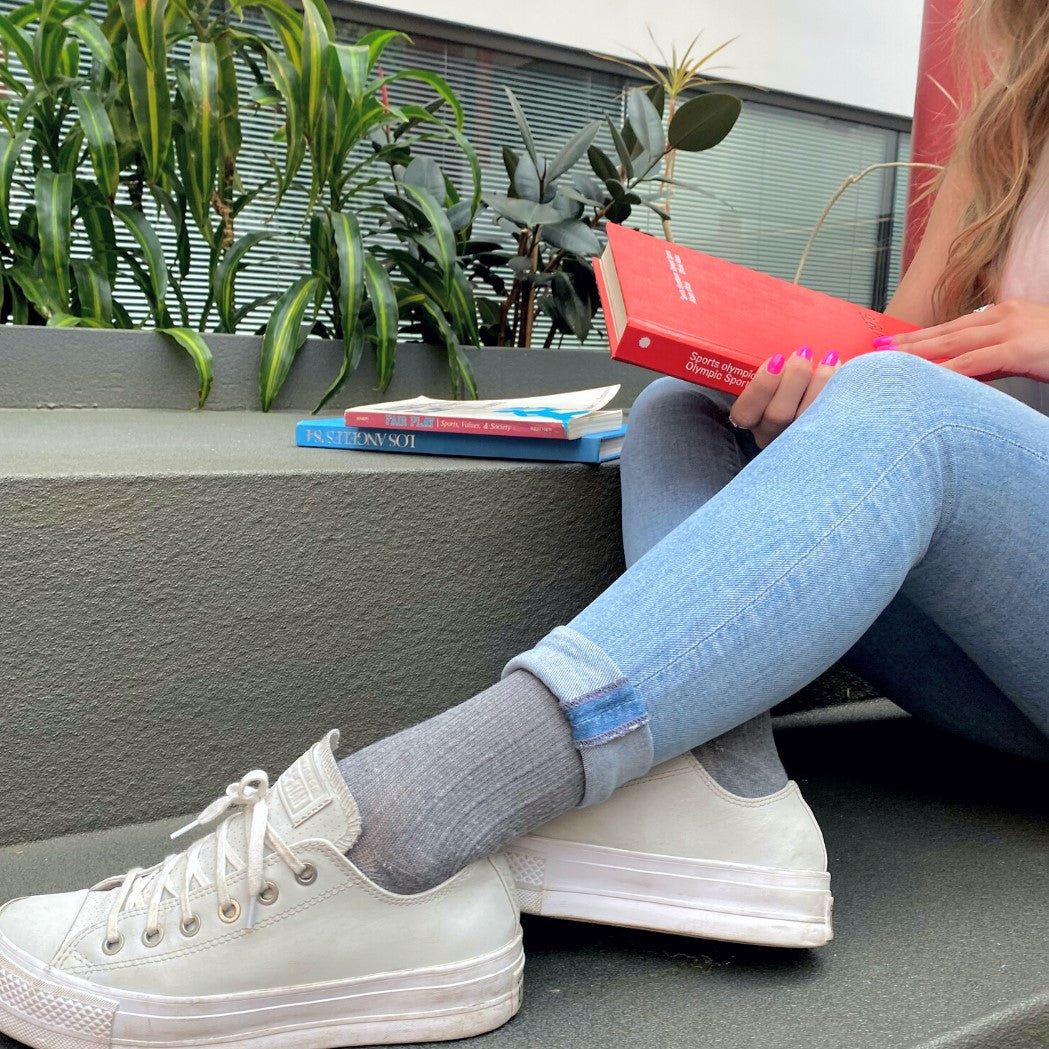 Young woman wearing sigvaris light grey linen compression socks with rolled-up skinny jeans and white sneakers, reading a book outdoors.
