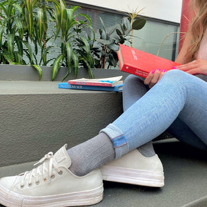 Young woman wearing sigvaris light grey linen compression socks with rolled-up skinny jeans and white sneakers, reading a book outdoors.