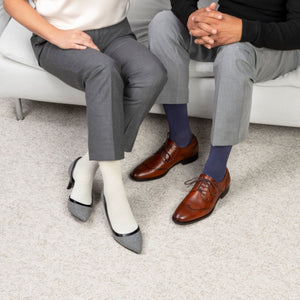 Two models in an office setting sitting on a couch. Left model is a female wearing the sigvaris bamboo sompression sock in Ivory, while the right model is a male, wearing the dusk violet color instead.