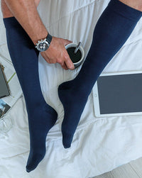 man sitting on edge of the bed with coffee wearing navy siglaris essential cotton compression socks