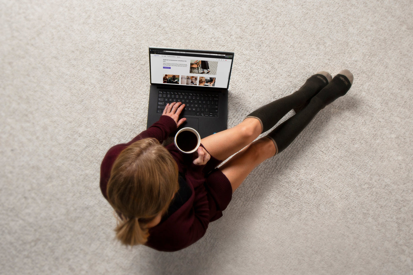 woman wearing the sigvaris merino outdoor knee high compression sock in olive, while working on a laptop and drinking coffee.